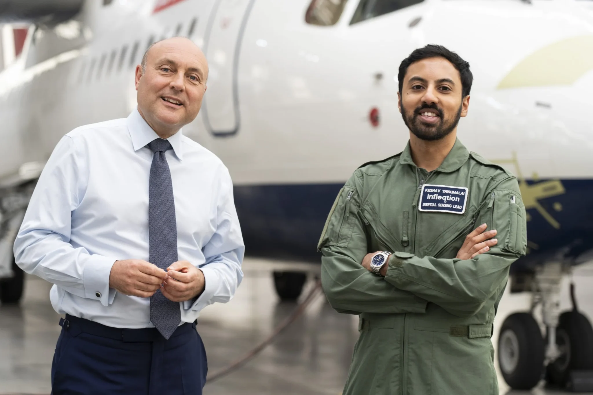 Two men pose in front of a plane, one of them wearing an Infleqtion branded flight suit.